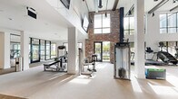 Located at 881 W Warren Ave in Longwood, Florida, this single-floor fitness center offers a generously sized, well-appointed workout facility that combines industrial-chic aesthetics with modern functionality. The space is defined by its dramatic double-height ceilings, exposed brick accent walls, and large black-framed windows that flood the interior with natural light and provide views of the surrounding outdoor area, including a glimpse of the pool beyond.

The main workout floor is thoughtfully organized into dedicated zones. The cardio area features a row of treadmills, ellipticals, and stair climbers aligned along the windowed wall, each equipped with individual screens for entertainment. Overhead, wall-mounted flat-screen TVs provide additional viewing options throughout the space.

The strength training section is equally well-equipped, housing a full rack of dumbbells, adjustable benches, multiple Precor cable and resistance machines, and a functional training zone with TRX straps, kettlebells, and medicine balls neatly organized on dedicated storage racks. A secondary, lower-ceilinged area offers additional selectorized machines in a quieter setting, complemented by floor-to-ceiling mirrors.

Exposed ductwork, track lighting, and decorative geometric wall art contribute to the space's contemporary industrial character. The light-toned carpet throughout keeps the environment clean and professional, making this a comprehensive and visually striking fitness facility.