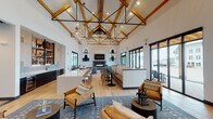 This single-story community clubhouse showcases a striking modern farmhouse aesthetic defined by its dramatically vaulted ceiling, where natural wood beams intersect with black steel truss framework beneath white shiplap panels. Pendant globe lights and circular chandeliers hang at varying heights, creating a layered lighting scheme that complements the warm light hardwood flooring throughout.

The open-concept main area integrates multiple functional zones seamlessly. A generously sized wet bar and kitchen area features a large white quartz island with bar seating, two-tone cabinetry pairing light wood uppers with dark lower cabinets, a pale blue subway tile backsplash, open display shelving with black metal framing, a stainless steel dishwasher, and a beverage refrigerator.

The social lounge area is anchored by a linear gas fireplace flanked by wall-mounted sconces and bold oversized artwork. Multiple seating arrangements—including leather chairs, plush sectional sofas, and built-in bench seating—define distinct gathering areas.

Floor-to-ceiling black-framed glass doors and windows on the perimeter flood the interior with natural light and provide direct access and views to an outdoor patio and surrounding community.