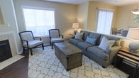 Located at 2221 Hyde Dr in Greenville, North Carolina, this single-story home offers a comfortable and well-appointed living environment. The living room serves as the heart of the space, showcasing a warm, neutral color palette with beige walls that create an inviting and relaxed atmosphere. Large windows dressed with white blinds and vertical shades allow generous amounts of natural light to fill the room, enhancing its open and airy feel. The living area is thoughtfully arranged with a plush gray sofa accented by decorative pillows, complemented by a pair of classic upholstered accent chairs with a subtle patterned fabric. A rustic wooden storage trunk coffee table anchors the seating arrangement atop a patterned area rug, adding a touch of character and practicality to the space. Soft ambient lighting is provided by a stylish table lamp, contributing to the room's warm and welcoming ambiance. The overall design of this home balances comfort and functionality, making it an ideal space for both relaxing and entertaining. The cohesive aesthetic throughout reflects a thoughtful approach to interior design, with attention to detail evident in every element of the living area.