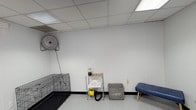 This single-floor pet grooming facility is purpose-built for professional pet care services, offering a clean, well-organized environment across its interconnected rooms. The space features crisp white paneled walls, drop tile ceilings with integrated fluorescent lighting, and durable speckled flooring throughout — all chosen for ease of maintenance and a hygienic working environment.

The grooming area is equipped with a stainless steel elevated bathing tub featuring built-in restraint loops and a handheld sprayer, alongside a raised grooming table with an adjustable grooming arm. These professional-grade fixtures are well-positioned to allow groomers to work efficiently and safely. A wall-mounted industrial fan provides additional ventilation and climate control, ensuring a comfortable atmosphere for both animals and staff.

A dedicated support area houses a wire kennel, a laundry basket for towels and linens, a multi-tier utility shelving unit stocked with grooming supplies, a handheld pet dryer, and a padded bench — offering practical staging space between grooming sessions. An interior window provides sightlines between rooms, enhancing supervision and workflow. Additional storage is available via a tall cabinet. The facility is thoughtfully laid out to support a smooth, professional grooming operation from intake to finish.