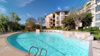 This outdoor amenity space offers a resort-like atmosphere centered around a generously sized swimming pool with crystal-clear blue water and stone tile edging. The pool area is well-equipped with lounge seating and shaded umbrella tables, providing comfortable spaces for relaxation. Adjacent to the pool, a bubbling spa with surrounding lounge chairs offers a dedicated area for unwinding. The grounds are thoughtfully landscaped with manicured hedgerows, ornamental ferns, sago palms, and mature trees that provide both shade and a sense of natural seclusion. Concrete walkways wind through the greenery, connecting the various outdoor zones in an intuitive and accessible manner. Wrought iron fencing borders portions of the space, adding a classic architectural detail that complements the multi-story building backdrop. The building itself features a warm stucco exterior, large grid-pane windows, and private balconies on each floor, contributing an elegant residential character to the outdoor environment. The overall ambiance is one of a well-maintained, upscale communal retreat, where lush greenery, open sky, and refined design elements come together to create an inviting and serene outdoor living experience.