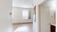 Located at 301 W Main Ave in the heart of downtown Spokane, Washington, this compact single-floor unit offers an efficient and functional layout comprising a kitchen, living room, bathroom, and closet. Light wood laminate flooring runs throughout the space, providing a clean and cohesive look that enhances the sense of openness.

The living room is bright and airy, featuring windows fitted with horizontal blinds that allow natural light to filter in while offering privacy. A baseboard heating system runs along the exterior walls, and a window-mounted air conditioning unit ensures year-round comfort. The walls are finished in a neutral white, complemented by a textured ceiling.

The kitchen is equipped with warm-toned wood cabinetry, both upper and lower, along with a white refrigerator, electric range, and a single-basin sink set into a countertop with a bold orange finish — a distinctive design element. A window in the kitchen brings in additional natural light.

The bathroom features a full tub-and-shower combination with floor-to-ceiling tile surround in a neutral beige tone, a toilet, and a vanity with a mirror. The tile work and fixtures are clean and functional.

A dedicated closet provides valuable storage in this efficiently designed space, making the most of every square foot in this centrally located Spokane unit.