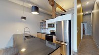 This single-floor space blends contemporary design with industrial character in a seamless and visually striking way. The kitchen serves as a central highlight, showcasing a large island topped with a rich, dark granite countertop that provides generous workspace and a bold aesthetic anchor. Light wood cabinetry lines the lower units, complemented by open metal-framed shelving above, creating a layered, modern storage solution. A white subway tile backsplash adds a clean, classic contrast to the darker tones throughout. Stainless steel appliances, including a full-size refrigerator, electric range, and over-the-range microwave, lend a polished, professional feel to the culinary space. The kitchen is completed by a sleek, industrial-style pull-down faucet set into the island sink. Overhead, exposed wooden beams cross the ceiling, adding warmth and a nod to the building's heritage character. Pendant lighting with matte black cylindrical shades hangs at varying heights, providing focused task lighting while enhancing the industrial-modern ambiance. Exposed ductwork and high ceilings contribute to the loft-like atmosphere, making the space feel open and expansive throughout.