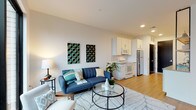 This single-floor unit showcases a contemporary open-concept layout where the living area and kitchen flow seamlessly together. The living space is bright and airy, anchored by large sliding glass doors that open onto a private balcony offering elevated urban views of the surrounding cityscape. Warm hardwood-style flooring runs throughout, complementing the clean, neutral palette of the walls and ceilings with recessed lighting.

The kitchen is well-appointed with white cabinetry, grey countertops, a subway tile backsplash, and a mix of white and stainless steel appliances. Open wood-bracketed shelving adds a modern touch alongside ample upper and lower cabinet storage. A deep single-basin sink and dishwasher complete the functional layout.

The bedroom is a quiet retreat with a tall, narrow window framing city views, allowing natural light while maintaining privacy. The bathroom is finished in a crisp all-white palette with grey countertops, a vanity with under-cabinet storage, and a full bathtub. A notable convenience is the stacked washer and dryer unit located directly adjacent to the bathroom, offering practical in-unit laundry. A bold dark-painted entry door adds a striking design accent at the unit's entrance.