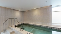 This tranquil indoor spa space is designed with relaxation and wellness in mind. At its centerpiece is a built-in pool or plunge tub, clearly marked with depth indicators and equipped with stainless steel handrails for easy entry and exit. The pool is framed by a mosaic tile border in neutral tones, adding a refined decorative detail that complements the overall aesthetic.

The walls are clad in large-format, light-toned tile panels with a subtle linear texture, creating a clean and sophisticated backdrop. A standout feature is the decorative accent wall panel, showcasing an intricate mosaic or dimensional tile work that serves as a focal point and adds visual depth to the space.

Illumination is provided by a combination of recessed ceiling lights and natural light streaming in through a large window, which brightens the room and contributes to an open, airy atmosphere. The smooth white ceiling enhances the sense of height and spaciousness. The flooring continues the neutral, spa-like palette with light-toned tile.

An adjacent corridor visible from the main area suggests additional rooms or amenities beyond, hinting at a broader wellness facility. The overall design is cohesive, understated, and elegant, offering a calming environment well-suited for therapeutic or leisure use.