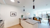 This contemporary single-floor residence is designed with a clean, modern aesthetic and thoughtful attention to detail throughout. The open-concept kitchen flows seamlessly into the living and dining areas, creating a cohesive and functional space. The kitchen features white flat-panel cabinetry, grey countertops, stainless steel appliances including a refrigerator, microwave, and range, as well as a subway tile backsplash. A peninsula with bar seating and a built-in sink serves as both a functional workspace and a casual dining spot, illuminated by a pair of sleek black cylindrical pendant lights overhead. Light hardwood flooring runs throughout, adding warmth to the bright, white-walled interior. The living area is anchored by a plush sectional sofa and benefits from generous natural light through large windows that frame views of the surrounding cityscape. The bedroom is a serene retreat, featuring a teal upholstered headboard, ample natural light, and an adjacent bathroom with white subway tile, a glass-enclosed shower, and clean chrome fixtures. A walk-in closet with hanging storage connects the bedroom and bath. The residence's modern barn-style door, recessed lighting, and curated wall art complete the polished, contemporary feel of this well-designed space.