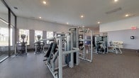 This fitness center is a comprehensively outfitted facility designed to support a full range of workout routines. The space is organized into distinct functional zones, all finished with durable rubber flooring and illuminated by recessed ceiling lighting throughout. The main strength training area features an extensive selection of Matrix-brand selectorized machines, a multi-station cable system, adjustable benches, and a well-stocked dumbbell rack spanning a broad weight range, complemented by full-length wall mirrors that enhance the sense of openness. A dedicated cardio zone along the windowed wall houses multiple Matrix treadmills, positioned to take advantage of natural light and exterior views. A separate spin studio, enclosed by floor-to-ceiling glass walls and doors, contains several stationary cycling bikes alongside a wall-mounted television, creating a semi-private environment ideal for cycling workouts. The entry area transitions smoothly into the fitness floor and includes an automated package locker system with wood-look flooring, adding a practical amenity to the facility. Elevated ceilings contribute to an airy, open ambiance throughout the space.