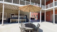 This single-story courtyard-style property combines inviting outdoor and well-appointed interior spaces. The central courtyard is an open-air gathering area, partially shaded by a pergola-style overhead structure and featuring ornate cast-iron patio furniture beneath a large market umbrella. A long counter or bar surface lines one side of the courtyard, providing a functional communal area. Upper-level balconies with white railings overlook the space, adding a layered architectural character, while surrounding terracotta-toned exterior walls lend a warm, cohesive aesthetic.

The interior units feature open living areas with smooth, neutral-toned walls, dark hardwood-style flooring, and ceiling fans that contribute to a comfortable, airy feel. Large windows allow natural light to fill the living spaces, which connect seamlessly to adjacent rooms. Kitchens are thoughtfully equipped with rich dark-wood cabinetry, stainless steel appliances including a refrigerator, dishwasher, and range, as well as ample counter space and a double basin sink. Hallways feature dark tile flooring and crisp white panel doors, providing access to closets and bathrooms. Overall, the space balances functional interior living with a welcoming communal outdoor environment.
