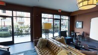 This sophisticated single-floor lounge area exudes a contemporary urban ambiance, thoughtfully designed to blend comfort with modern style. The space is anchored by a richly tufted caramel leather sofa adorned with plush, textured throw pillows that add a warm, inviting touch. Deep espresso-brown walls create a bold, intimate atmosphere that is elegantly balanced by the abundance of natural light streaming through large, floor-to-ceiling windows and glass doors. These expansive windows frame views of a well-appointed outdoor patio area, complete with seating, and offer glimpses of the surrounding urban streetscape. A striking table lamp with a mosaic-style base and a warm-toned shade serves as a notable decorative accent. Sleek, dark upholstered seating and a polished side table round out the lounge area, contributing to the space's refined aesthetic. The flooring transitions from a light-toned tile near the entry and patio access points to a softly hued area rug beneath the main seating arrangement. Artwork adorns the walls, adding a curated, gallery-like quality. Overall, the space strikes a balance between urban sophistication and approachable comfort, making it well-suited as a communal gathering or hospitality lounge.