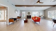 Located at 7325 Goldenpointe Blvd in Orlando, FL, this single-story clubhouse offers a well-appointed community gathering space designed to serve residents with both leisure and functional amenities. The main lounge is a highlight of the interior, featuring vaulted ceilings with striking exposed wood beams, light wood-style flooring, recessed lighting, and a modern ceiling fan. Large windows flood the space with natural light and offer views of the surrounding greenery, creating a bright and welcoming atmosphere. The lounge is open and generously proportioned, seamlessly connecting to a dedicated workspace area complete with a desk and seating — ideal for remote work or study. A welcoming entry foyer with a reception counter, stacked stone accent wall, and elegant French doors with glass panel detailing sets the tone upon arrival. The fitness center is well-equipped with treadmills, an elliptical, a stationary bike, free weights, exercise balls, and wall mirrors, all set on durable rubber flooring. A full bathroom is also available on-site. Outdoors, a large swimming pool surrounded by lounge chairs, covered gazebos, and lush tropical landscaping completes this comprehensive community amenity space.