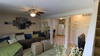 This two-floor residence features an open-concept main level that seamlessly connects the living room, dining area, and kitchen. The living room is a comfortable, well-appointed space anchored by a ceiling fan with an integrated light fixture and a textured ceiling. The room accommodates generous seating, including a large sectional sofa accented with decorative pillows, complemented by woven ottoman coffee tables and lush indoor plants that add natural character to the space.

Structural columns define the transition between the living area and the rest of the main level, preserving an airy, open feel while providing architectural interest. The dining area sits just beyond, furnished with a dark bistro-style table and chairs. The kitchen, visible from the living room, features light wood cabinetry, a white refrigerator, and light-toned flooring, creating a bright and functional cooking space. A decorative "EAT" sign adds a casual, welcoming touch.

The flooring transitions from plush carpet in the living area to smooth hard-surface flooring in the kitchen and dining zones. Warm, ambient lighting throughout the main level contributes to an inviting and cohesive atmosphere across the shared spaces.