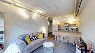 This single-floor space offers an efficient and functional open-concept layout that seamlessly combines the living and kitchen areas into one cohesive environment. The living area is anchored by a comfortable neutral-toned sofa accented with colorful throw pillows in blue and yellow, paired with a minimalist wire side table that adds a contemporary touch. Overhead, a track lighting system runs the length of the ceiling, providing flexible and well-distributed illumination throughout the space while contributing to its modern, urban aesthetic. The kitchen is positioned directly adjacent to the living area and features light-wood upper cabinetry, a white refrigerator, a range, and a microwave. A peninsula counter serves as both a prep space and a casual dining area, lined with sleek metal bar stools that reinforce the industrial-modern design sensibility. The flooring transitions subtly between the two zones, further defining each area while maintaining an open, airy feel. Exposed structural ceiling elements add an industrial character to the space, giving it a loft-like quality. A hallway visible in the background suggests additional rooms or storage beyond the main living area. Overall, the space is practical, well-lit, and thoughtfully arranged to maximize functionality.