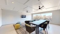 This covered rooftop lounge offers a well-appointed communal retreat designed for both relaxation and collaboration. The space is defined by its crisp white shiplap walls and a geometric coffered ceiling fitted with two large modern ceiling fans, ensuring comfort throughout the year. Recessed lighting complements the abundant natural light that floods in through a wide, panoramic window framing impressive city skyline views and a railed balcony beyond.

The layout is thoughtfully organized to accommodate multiple uses. A long high-top dining table with woven rope bar chairs provides ample seating for group gatherings or working sessions, while a distinct lounge area features an eclectic mix of rounded accent chairs, sculptural ottomans in teal and yellow, and a patterned area rug that anchors the space with warmth and visual interest. A wall-mounted flat-screen TV is positioned prominently on the shiplap feature wall, making the space ideal for presentations or casual viewing.

Decorative touches, including a botanical wall art piece and vibrant pops of color throughout the furnishings, lend the space a contemporary, energetic character. The combination of indoor comfort and open sightlines to the urban landscape makes this rooftop lounge a versatile and inviting environment.