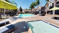This inviting outdoor amenity space centers around a generously sized swimming pool with crystal-clear blue water, complemented by an adjacent spa for relaxation. The pool deck is finished in warm-toned stone pavers, providing ample space for lounging and recreation under the California sun. Tall, mature palm trees and shade trees line the perimeter, lending a tropical, resort-like ambiance to the area. A series of sleek metal pergola structures run alongside the pool, each sheltering contemporary light-grey outdoor dining tables and chairs — ideal for shaded gatherings or casual outdoor meals. The landscaping throughout features a thoughtful mix of drought-tolerant plants, decorative rock beds, and trimmed greenery framed by stone retaining walls and black iron fencing. The backdrop reveals a striking mountain range that rises above the rooftops of the surrounding residential buildings, adding a dramatic natural element to the setting. The overall design of the space balances functionality and aesthetics, offering residents a well-appointed outdoor retreat with multiple seating zones, water features, and scenic views that make it suitable for both quiet relaxation and social gatherings throughout the year.