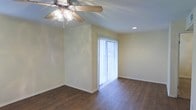 This single-story space showcases a clean, functional design highlighted by vaulted ceilings that lend an open, airy quality to the interior. The main living area is finished with rich, dark hardwood flooring that provides a warm contrast against the light, neutral-toned walls. A ceiling fan with an integrated light fixture is centrally mounted, offering both comfort and ambient lighting. A sliding glass door dressed with vertical blinds serves as a prominent architectural feature, drawing in generous natural light while providing access to the outdoors. Recessed lighting adds a modern touch to complement the overall brightness of the space. White baseboards frame the perimeter of the room, adding a crisp, finished look. The open layout and vaulted ceiling together create a sense of spaciousness, making the area feel welcoming and versatile for a variety of uses.