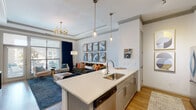 This single-floor residence is thoughtfully designed with an open-concept layout that seamlessly connects the kitchen and living areas. The kitchen is a standout feature, showcasing gray shaker-style cabinetry with crown molding detail, quartz countertops, a subway tile backsplash, and stainless steel appliances including a range, microwave, and dishwasher. A central island with a stainless steel sink and pendant lighting above provides additional prep space and casual seating with room for bar stools.

The living area benefits from large windows and a glass door that flood the space with natural light and offer outdoor views. A tray ceiling with crown molding adds architectural depth, while a decorative chandelier enhances the refined ambiance. Wood-style flooring runs throughout the main living spaces, transitioning to plush carpet in the bedroom.

The bedroom features a dedicated accent wall, a ceiling fan, and generously sized windows framed by floor-length curtains, creating a serene, private retreat. The bathroom includes a full soaking tub with a tiled surround and clean white finishes. Throughout the residence, an elegant and cohesive design aesthetic is maintained, blending contemporary style with comfortable, functional living.