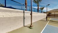 Located at 4550 S 67th Ave in Phoenix, Arizona, this outdoor recreational facility features two dedicated pickleball courts designed for year-round play in the Arizona sunshine. The courts are finished in a dual-tone blue and gray surface with crisp white line markings and regulation nets, providing a professional playing experience. Each court is equipped with sturdy net posts, and the facility is outfitted with multiple tall stadium-style light poles to support evening play. The courts are enclosed by a combination of chain-link fencing and solid concrete block perimeter walls, ensuring a contained and secure playing environment. Tall, mature palm trees line the perimeter, adding a distinctly Southwest aesthetic and providing partial natural shade. An adjacent single-story building with a tile roof, stone-accented façade, and a gated entry offers convenient access and support facilities for players. The overall layout is open-air, taking full advantage of the abundant natural light and expansive Arizona sky. The thoughtful design balances functionality with visual appeal, making this a well-appointed recreational amenity suited for players of all skill levels.
