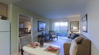 This single-floor residence offers an open and cohesive layout connecting the main living area to the bedroom. The living room is finished with soft sage green walls and plush wall-to-wall carpeting, creating a warm and comfortable atmosphere. A sliding glass door fills the space with natural light and provides direct access to a private patio, where lush greenery is visible, enhancing the indoor-outdoor connection.

The living area is thoughtfully arranged with ample room for a sofa, accent chairs, and a central coffee table, making it well-suited for both relaxing and entertaining. A framed wall hanging and a console table with decorative accents add character to the space. A table lamp provides warm supplemental lighting alongside the abundant natural light from the patio door.

An open doorway connects the living room to the bedroom, which features a dark wood bed frame and coordinating furnishings. The consistent use of carpeting and the sage green color palette throughout creates a unified aesthetic. A mirror near the living area further enhances the sense of openness, making the overall space feel bright and inviting.
