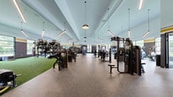 Located at 201 Munson Street in New Haven, Connecticut, this expansive fitness center is a comprehensively equipped exercise facility spanning a single, open floor. The space is divided into several distinct training zones, each thoughtfully designed to support a full range of workout modalities.

The main strength and resistance area features a wide selection of selectorized machines, cable systems, and free-weight benches, all set on durable speckled rubber flooring. Soaring ceilings throughout create an open, airy atmosphere, while a combination of linear pendant lighting and recessed fixtures ensures excellent visibility across the entire floor. Bold navy blue walls accented with gold and white striping lend the space a dynamic, athletic aesthetic.

A dedicated functional training zone is outfitted with artificial turf, providing an ideal surface for agility drills, medicine ball work, battle ropes, and tire training. The cardio section is lined with a row of treadmills and stationary equipment positioned in front of large picture windows that frame outdoor views, filling the area with natural light.

A separate studio space, distinguished by its soaring arched windows, floods the room with abundant daylight and accommodates cycling machines and group fitness equipment. Throughout the facility, floor-to-ceiling windows and generous mirror placements amplify the sense of space and connectivity to the outdoors, creating an energizing environment conducive to peak performance.