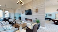 This well-appointed clubroom delivers a refined yet welcoming atmosphere, thoughtfully designed for both relaxation and socializing. The focal point of the space is a striking floor-to-ceiling textured accent wall finished in soft grey tones, which houses a sleek linear electric fireplace and a wall-mounted flat-screen television — a combination that anchors the room with both warmth and modern functionality. Vaulted ceilings amplify the sense of openness, while a classic chandelier and decorative wall sconces cast a warm, layered glow throughout the room. The seating area is furnished with a curated mix of wicker and upholstered chairs arranged around a woven coffee table, evoking a relaxed coastal aesthetic. Soft neutral carpeting and light-toned flooring complement the cool blue accent walls visible in the adjoining area, creating a cohesive and calming color palette. Potted greenery and coastal décor accents — including coral and sea-inspired objects — add natural texture and character to the space. An adjacent room, visible through the open layout, provides additional seating, further extending the clubroom's capacity for gathering and entertaining. Overall, the space strikes a balance between casual comfort and polished design.