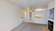 This single-floor unit offers a practical and efficient layout designed to maximize its compact footprint. The main living area features neutral-toned carpeting and light-colored walls that create a bright, open feel. A ceiling fan is centrally positioned overhead, providing both air circulation and ambient lighting to the space.

A large walk-in closet with built-in shelving is accessible directly from the living area, offering organized storage solutions. Natural light enters through a window adjacent to the closet, adding brightness to the interior.

The kitchen is positioned along one side of the unit and is accessible via a pass-through counter that opens to the living space, creating a connected flow between the two areas. The kitchen includes a stovetop, sink, and upper cabinetry finished in white, contributing to a clean, uniform aesthetic. An under-cabinet light fixture provides functional task lighting.

A wall-mounted heating unit is also present in the living area. The overall design is straightforward and functional, with a neutral color palette throughout that lends itself well to a variety of furnishing styles.