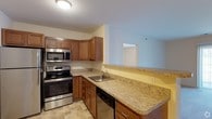 This single-floor residence offers a practical and comfortable layout designed to maximize everyday livability. The kitchen is a standout feature, showcasing rich medium-toned wood cabinetry that provides generous storage space above and below the countertops. The light granite-style countertops extend throughout the kitchen, offering ample prep and workspace. Stainless steel appliances — including an electric range, over-the-range microwave, and dishwasher — lend a modern, cohesive look to the space. A stainless steel undermount sink is neatly positioned along the cabinetry, completing the kitchen's functional design. The kitchen flows openly toward the adjacent living area through a pass-through opening, enhancing the sense of connectivity between spaces. Light tile flooring runs throughout, contributing to a clean and cohesive aesthetic. The walls are painted in soft, neutral tones — a combination of warm yellow and cool grey — that create a welcoming ambiance. Overall, the layout is thoughtfully designed to balance utility and comfort, making it a practical space suited for everyday living.