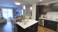 This well-appointed single-floor residence offers an open-concept design that integrates the kitchen, dining, and living areas into one cohesive and functional space. The modern kitchen is a standout feature, showcasing rich dark cabinetry that contrasts beautifully with crisp white countertops. A central island with pendant lighting above serves as both a prep space and a gathering point, adding to the kitchen's practicality and visual appeal. Stainless steel appliances, including a dishwasher and refrigerator, complete the sleek culinary setup. Warm hardwood-style flooring runs throughout, lending a sense of continuity and warmth across the open floor plan. The dining area is thoughtfully positioned adjacent to the kitchen, accommodating a full dining set ideal for entertaining or everyday meals. Beyond the dining space, the living room provides a comfortable retreat, complete with a mounted television and natural light filtering in through window treatments. Framed landscape artwork adorns the walls, adding a curated, elevated feel to the interior. The overall ambiance is modern yet inviting, with a neutral color palette, tasteful décor, and a well-balanced mix of form and function throughout.