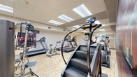 Located at 3021 Maple Valley Dr in Madison, WI, this fitness center is a comprehensive single-floor facility divided into two dedicated exercise rooms. The space is designed to accommodate a full range of workout needs, combining cardio and strength training areas in a clean, functional layout.

The primary cardio room features light hardwood-style flooring and a drop tile ceiling with recessed skylight panels that provide bright, even illumination throughout. This room is well-stocked with treadmills, elliptical trainers, a stationary bike, and a StairMaster, along with a wall-mounted television for entertainment during workouts. A water cooler station is conveniently positioned within the room.

A striking floor-to-ceiling mural depicting the Madison city skyline at sunset serves as a bold focal point, adding a vibrant splash of color and local character to the space. The artwork spans an entire accent wall, creating an energizing atmosphere for users.

The second exercise room transitions to a darker rubber flooring, signaling its purpose as a dedicated strength training area. This room is outfitted with free weights, barbells, a weight rack, adjustable benches, a cable pulley machine, and a step platform. Large wall mirrors enhance the sense of space and allow users to monitor their form. Both rooms are well-lit and maintain a clean, functional aesthetic throughout.