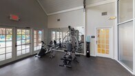 This single-floor fitness center is defined by its striking architectural character, most notably the soaring vaulted ceiling with exposed wooden trusses that lend the space a warm yet open industrial aesthetic. Abundant natural light filters in through multiple sets of French doors and large frosted glass panel walls, creating a bright and airy workout environment throughout.

The space is organized into distinct functional zones. A cardio area along one wall features two treadmills and an elliptical trainer, all positioned in front of French doors that overlook an outdoor pool area, providing an engaging view during workouts. A strength training section includes a multi-function cable machine, a weight bench, and a set of free weights with a rack, complemented by a large wall mirror that visually expands the room and aids in form correction. A dedicated stretching and functional fitness area houses a medicine ball tower with a colorful assortment of weighted balls, along with ankle weights stored nearby.

Additional amenities include a water cooler for hydration and a rubber flooring surface throughout that provides safety and comfort underfoot. The space is accessed through a glass-paneled entry door and connects to an adjoining room via a French door, rounding out a comprehensive and inviting fitness facility.