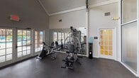 This single-floor fitness center is defined by its striking architectural character, most notably the soaring vaulted ceiling with exposed wooden trusses that lend the space a warm yet open industrial aesthetic. Abundant natural light filters in through multiple sets of French doors and large frosted glass panel walls, creating a bright and airy workout environment throughout.

The space is organized into distinct functional zones. A cardio area along one wall features two treadmills and an elliptical trainer, all positioned in front of French doors that overlook an outdoor pool area, providing an engaging view during workouts. A strength training section includes a multi-function cable machine, a weight bench, and a set of free weights with a rack, complemented by a large wall mirror that visually expands the room and aids in form correction. A dedicated stretching and functional fitness area houses a medicine ball tower with a colorful assortment of weighted balls, along with ankle weights stored nearby.

Additional amenities include a water cooler for hydration and a rubber flooring surface throughout that provides safety and comfort underfoot. The space is accessed through a glass-paneled entry door and connects to an adjoining room via a French door, rounding out a comprehensive and inviting fitness facility.