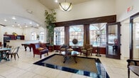 This single-floor clubhouse facility offers a thoughtfully designed interior that blends elegance with functionality across multiple distinct areas. The grand entrance foyer sets an impressive tone with soaring ceilings, gleaming tile flooring with dark inlay accents, a decorative round table centerpiece, an ornate chandelier, and stately double doors with wrought iron detailing that invite natural light inside.

The main lounge is a standout space, featuring dramatic vaulted ceilings, large leaded-glass windows overlooking an outdoor pool area, rich leather seating, tall indoor plants, and a sunken conversation area defined by contrasting tile inlay and an area rug. Floor lamps and pendant lighting add warmth throughout.

A dedicated fitness center is bright and airy, with arched windows, high ceilings, a ceiling fan, wood-look flooring, and a full complement of equipment including treadmills, ellipticals, cable machines, and a free-weight rack.

The billiards room provides a relaxed recreational atmosphere with large windows, tile flooring, a full-size pool table, mounted television, and an adjacent kitchenette bar area. A cozy café nook nearby offers booth-style seating, bistro tables, and charming décor details.

A private conference or meeting room features wood-paneled wainscoting, wood-look floors, a long table with mixed seating, and framed artwork — ideal for professional or community gatherings.