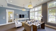 Located at 14411 Calloway Gap Rd in Raleigh, NC, this single-floor space offers a beautifully curated interior that blends comfort and refined style. The living area immediately draws attention with its coastal-inspired shiplap accent wall finished in a calming steel blue, creating a warm yet sophisticated atmosphere. Elevated ceilings enhance the sense of openness, while a large multi-pane window floods the room with natural light and frames lush greenery views of the surrounding landscape. Stylish pendant and drum light fixtures with brass accents add warmth and a touch of elegance to the space. The seating arrangement is thoughtfully composed, featuring tufted upholstered sofas, accent chairs in crisp white, and throw pillows in complementary blue and neutral tones. A white media console serves as a functional and decorative focal point along the accent wall, complemented by coordinating table lamps. The space also incorporates a wooden side table and desk area, adding a practical yet tasteful touch. Rich, patterned carpeting grounds the room while harmonizing with the cool blue-gray wall tones. Overall, the space exudes a polished, inviting ambiance suited for both relaxed living and comfortable entertaining.
