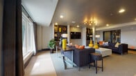 This single-floor communal space is thoughtfully designed to accommodate a variety of social and functional needs, seamlessly integrating a lounge area, dining zone, and a well-appointed kitchen. The open layout encourages fluid movement between each area, unified by light wood-toned tile flooring and neutral wall tones throughout.

The lounge area is anchored by plush dark sofas accented with vibrant orange and patterned throw pillows, centered around a substantial stone-finish coffee table. A wall-mounted flat-screen television is set against a textured brick-style feature wall, flanked by built-in shelving displaying curated décor. An eye-catching multi-arm sputnik chandelier provides warm, ambient lighting overhead, complemented by recessed ceiling fixtures.

The dining area transitions naturally from the lounge, featuring both a large communal table with upholstered chairs and an extended bar-height counter with tall seating—ideal for casual gatherings. Sculptural pendant light fixtures in brushed gold tones hang above, adding a refined touch.

The kitchen runs along one side of the space, offering generous counter space with polished stone surfaces, dark cabinetry, and modern fixtures. A glass-paneled entry corridor with light wood flooring provides a clean, accessible entrance, with views into an adjacent fitness area. Large windows allow natural light to filter in, with drapes framing views toward an outdoor terrace.