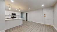 This thoughtfully renovated single-floor space presents a clean, modern aesthetic anchored by a cohesive neutral palette and light-toned luxury vinyl plank flooring that flows seamlessly throughout. The open-concept kitchen is a standout feature, showcasing crisp white shaker cabinetry with matte black hardware, a herringbone-patterned tile backsplash, and a full suite of stainless steel appliances including a gas range, built-in microwave, and dishwasher. A centered kitchen island with a bright white countertop provides additional workspace and serves as a natural divider between the kitchen and the generously sized living area. Recessed lighting and elegant pendant fixtures illuminate the space, enhancing its airy, welcoming feel. The living area offers ample room and is conveniently adjacent to a closet, providing practical storage. Two bedrooms continue the consistent design language, each featuring the same durable flooring and neutral grey walls, with windows that allow natural light to filter in. The full bathroom is finished with large-format grey tile flooring, a white vanity with a matte black faucet, a mirrored medicine cabinet, and a full soaking tub with shower — combining style and functionality in a well-appointed package.
