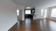 This single-floor space showcases an open-concept design that seamlessly connects the main living area to the kitchen. Rich, dark hardwood flooring extends throughout, creating a warm and cohesive aesthetic across the entire space.

The kitchen is well-appointed with dark cabinetry that contrasts sharply against light-colored countertops, likely granite or a similar stone surface. Stainless steel appliances, including a refrigerator, dishwasher, microwave, and range, provide a modern and functional cooking environment. A peninsula with pendant lighting serves as a natural divider between the kitchen and the open living area, offering additional counter space and a casual dining or workspace option.

Natural light is a defining feature of this space, with multiple windows fitted with white blinds allowing generous daylight to fill the interior. The white walls enhance this brightness, contributing to an airy and clean atmosphere throughout.

A doorway visible from the main area leads to an adjacent room, also benefiting from natural light, suggesting additional functional spaces connected to the open layout. The overall design is modern, neutral, and move-in ready.