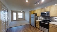This one-bedroom, one-bathroom unit offers an efficient and well-designed single-floor layout that maximizes its living space. The open-concept design seamlessly connects the kitchen and living areas, creating a cohesive and functional environment.

The kitchen is outfitted with stainless steel appliances, including a full-size refrigerator, gas range, and built-in microwave. Warm honey-toned cabinetry provides ample storage, while a mosaic tile backsplash adds a decorative accent above the light-colored countertops. Track lighting overhead ensures focused illumination for cooking and food preparation.

The adjacent living area transitions from the kitchen via hardwood-style flooring that extends throughout the main space. A ceiling fan with a light fixture provides both comfort and supplemental lighting. The living area is carpeted and benefits from natural light streaming through multiple windows, which also offer views to an adjoining balcony or patio space. Horizontal blinds offer privacy when needed.

Overall, the unit presents a clean, contemporary aesthetic with practical finishes suited for everyday living.