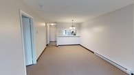 This single-floor residence offers a clean, functional layout with a neutral color palette that creates a bright and welcoming atmosphere throughout. The main living area is open and spacious, finished with wall-to-wall carpeting in a warm beige tone and complemented by crisp white walls that maximize the sense of light and space. A baseboard heating system runs along the perimeter, ensuring efficient climate control throughout the unit.

The kitchen is defined by a white peninsula counter that opens directly to the living area, creating a connected flow between the two spaces. Above the dining area, an elegant chandelier with multiple arms provides a tasteful focal point and warm ambient lighting. The kitchen itself features cabinetry and countertops visible through the pass-through opening, offering practical storage and workspace.

A hallway extending from the main living space leads to additional rooms, with tile flooring in the entryway transitioning from the carpeted living area. A mirrored sliding closet door near the entry adds depth and reflects light back into the room. The overall design is practical and move-in ready, with a neutral aesthetic that provides a versatile canvas for a variety of interior styles.