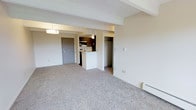 This single-floor unit offers a clean, functional layout designed to maximize everyday comfort. The main living area features neutral-toned carpeting that extends throughout, creating a warm and cohesive atmosphere. A baseboard heater along the exterior wall ensures year-round comfort, while the open floor plan allows for flexible furniture arrangements to suit a variety of lifestyles.

The kitchen is positioned at the heart of the unit and is defined by a raised breakfast bar that separates it from the living space. Dark wood cabinetry provides ample storage, complemented by a standard range and under-cabinet lighting that adds a practical warmth to the cooking area. The kitchen transitions smoothly into the adjoining space, making it well-suited for both daily use and casual entertaining.

A walk-in closet is conveniently located just off the main living area, offering generous storage space. The entry door, finished in a neutral gray, provides a clean and modern first impression upon arrival. Structural ceiling beams add subtle architectural character to the space. The overall palette of soft beiges and warm neutrals throughout lends the unit a calm, move-in-ready ambiance ideal for comfortable living.