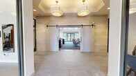 Located on Lighthouse Lane in East Lyme, Connecticut, this thoughtfully designed single-floor clubhouse-style space blends contemporary aesthetics with warm, inviting materials throughout. The entry foyer makes a strong first impression with large-format stone tile flooring, light wood-paneled walls, and a coffered ceiling fitted with elegant drum pendant fixtures. A pair of sliding barn doors open into the main lounge, establishing an easy transition between zones.

The central lounge is anchored by soaring vaulted ceilings with exposed dark timber trusses, creating dramatic architectural interest overhead. A linear fireplace with a wall-mounted television serves as the primary focal point, surrounded by stylish swivel chairs and mid-century-inspired furnishings arranged atop patterned blue carpeting. Floor-to-ceiling glass doors flood the space with natural light and provide direct views of the wooded landscape beyond an outdoor deck.

Adjacent to the lounge is a well-appointed bar and social area featuring a white quartz island with bar seating, pendant lighting, and a striking navy patterned tile backsplash. Wood-look tile flooring and a slatted wood ceiling add warmth to this gathering space. Additional seating nooks with textured wallcoverings, high-top tables, and intimate lounge chairs offer flexible areas for casual meetings or relaxation. A glass-walled fitness area is also visible, seamlessly integrated into the overall layout.