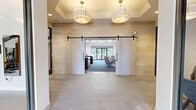 Located on Lighthouse Lane in East Lyme, Connecticut, this thoughtfully designed single-floor clubhouse-style space blends contemporary aesthetics with warm, inviting materials throughout. The entry foyer makes a strong first impression with large-format stone tile flooring, light wood-paneled walls, and a coffered ceiling fitted with elegant drum pendant fixtures. A pair of sliding barn doors open into the main lounge, establishing an easy transition between zones.

The central lounge is anchored by soaring vaulted ceilings with exposed dark timber trusses, creating dramatic architectural interest overhead. A linear fireplace with a wall-mounted television serves as the primary focal point, surrounded by stylish swivel chairs and mid-century-inspired furnishings arranged atop patterned blue carpeting. Floor-to-ceiling glass doors flood the space with natural light and provide direct views of the wooded landscape beyond an outdoor deck.

Adjacent to the lounge is a well-appointed bar and social area featuring a white quartz island with bar seating, pendant lighting, and a striking navy patterned tile backsplash. Wood-look tile flooring and a slatted wood ceiling add warmth to this gathering space. Additional seating nooks with textured wallcoverings, high-top tables, and intimate lounge chairs offer flexible areas for casual meetings or relaxation. A glass-walled fitness area is also visible, seamlessly integrated into the overall layout.