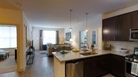 Located at 1 Kennedy Ave in Danbury, Connecticut, this single-floor residence showcases a thoughtfully designed open-concept layout that seamlessly connects the kitchen, dining, and living areas. The kitchen is a standout feature, boasting dark espresso cabinetry, gleaming granite countertops, a large center island with a stainless steel double sink, and pendant lighting that adds a warm, modern touch. Stainless steel appliances, including a dishwasher, complete the space. The open floor plan flows naturally into the living area, where rich hardwood-style flooring extends throughout, creating a cohesive and inviting ambiance. The living room is generously proportioned, bathed in natural light from large windows dressed with patterned curtains, offering a bright and welcoming atmosphere. Recessed lighting throughout enhances the clean, contemporary aesthetic. A glimpse into an adjoining room suggests additional well-appointed spaces beyond the main living area. The overall design balances functionality and style, making this residence an ideal space for both everyday living and entertaining.