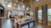 The Grand Parlor is a thoughtfully designed single-floor gathering space that blends rustic and contemporary elements to create a refined, welcoming atmosphere. The room's most commanding feature is its soaring ceiling height, accentuated by shiplap wall paneling and a striking rectangular candelabra-style chandelier that serves as a dramatic focal point. A floor-to-ceiling natural stone fireplace anchors one end of the space, crowned by a large circular mirror that reflects light throughout the room.

The parlor is organized into distinct yet open seating zones. The primary lounge area features neutral-toned sofas arranged around a central console table, adorned with sculptural décor including a notable white horse figurine. A built-in entertainment wall with dark wood shelving and cabinetry provides both functional storage and visual interest. Adjacent to this area, a dining zone with shiplap walls and multiple windows offers ample natural light and outdoor views.

Wide-plank hardwood flooring runs throughout, unifying the space, while recessed lighting and large windows ensure a bright, open ambiance. Neutral drapery panels frame the windows, complementing the warm, earthy tones of the overall palette. The Grand Parlor effectively balances elegance with comfort, making it a versatile and inviting communal space.