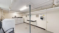 Located at 7 Laurel Dr in Scranton, PA, this dedicated community laundry facility offers a practical and functional space designed to accommodate multiple users efficiently. The room is well-lit by overhead fluorescent lighting and benefits from natural light through a multi-pane window, with views of the surrounding greenery outside. A wide exterior door provides direct access to an outdoor patio area, enhancing ventilation and ease of access. The facility is equipped with a row of front-loading washing machines and a set of commercial-grade dryers, numbered for easy identification. A sturdy folding table is positioned along one wall, providing ample surface space for sorting and folding laundry. A metal bench is centrally placed, offering a comfortable waiting area for users. The exposed pipe infrastructure along the ceiling gives the space an industrial character, while the light-colored walls and concrete floor contribute to a clean, utilitarian aesthetic. Safety features include a mounted fire extinguisher and an emergency access cabinet. A coin-operated payment system is conveniently installed on the wall. The space is thoughtfully organized to support a smooth and efficient laundry experience for all residents.
