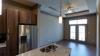 This single-floor residence offers a thoughtfully designed open-concept layout that seamlessly connects the kitchen and living areas. The kitchen is a standout feature, showcasing granite countertops with an undermount double sink, rich dark wood cabinetry, and stainless steel appliances, including a French door refrigerator. A pendant light fixture adds a refined focal point above the kitchen island, complementing the warm, contemporary aesthetic throughout.

The living area benefits from a striking tray ceiling detail, which adds architectural depth and dimension to the space. A ceiling fan with a light kit enhances both comfort and style. Rich dark hardwood flooring runs throughout, grounding the space with warmth and continuity. Neutral-toned walls in a cool gray-blue palette create a calm, versatile backdrop that suits a variety of décor styles.

Natural light flows generously into the space through a large entry door with transom window, offering a bright and welcoming ambiance. The overall design strikes a balance between modern practicality and classic elegance, with carefully chosen finishes and architectural details that elevate the everyday living experience. The open sightlines between the kitchen and main living area make this space feel expansive and ideal for both everyday living and entertaining.