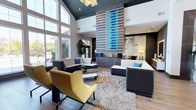 This single-floor space presents a striking contemporary lobby lounge designed to impress and welcome. The defining feature of the room is its soaring two-story ceiling finished in a deep teal-gray tone, which lends dramatic contrast against the bright white walls and abundant natural light streaming in through floor-to-ceiling windows along the perimeter. A bold, illuminated vertical feature wall serves as the room's centerpiece — constructed with layered wood slats and vivid blue LED lighting, it draws the eye immediately upon entry and anchors the entire space. The seating arrangement is thoughtfully curated, combining modular dark gray sofas, a crisp white sectional accented with blue throw pillows, and vibrant yellow accent chairs that introduce a pop of color and energy. A plush, textured area rug grounds the central seating cluster, adding warmth and depth to the wood-look flooring beneath. Lush indoor plants are positioned throughout, softening the modern aesthetic and bringing a touch of organic life to the interior. A contemporary chandelier overhead completes the refined look. The overall ambiance is polished yet approachable — balancing bold design choices with comfortable, functional furnishings ideal for a communal or hospitality setting.