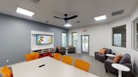 This single-floor multipurpose space is thoughtfully designed to support both collaborative work and casual gatherings. The room features a clean, contemporary aesthetic defined by white and light gray walls accented with a bold slate-blue feature wall, all set atop durable, neutral-toned carpet tiles that lend a professional yet comfortable feel throughout.

At the heart of the space sits a large white conference table surrounded by vibrant orange mesh rolling chairs, providing ample seating for group meetings or collaborative sessions. A wall-mounted flat-screen display on a sleek white media panel serves as the focal point for presentations, accompanied by a full-size whiteboard on the opposite wall for brainstorming and note-taking.

A dedicated lounge area offers a change of pace, furnished with two gray upholstered armchairs accented with orange patterned throw pillows and paired with modern white pedestal side tables — ideal for informal conversations or breakout discussions. An additional sofa rounds out the seating options near the display wall.

Natural light filters in through multiple windows fitted with horizontal blinds, creating a bright and open atmosphere. A striking black ceiling fan anchors the space overhead, complementing the recessed panel lighting. A glass-panel exterior door provides direct access to an outdoor area, further enhancing the sense of openness. Colorful accent décor, including vibrant floral artwork and bold orange sculptural elements, adds energy and personality to the overall environment.