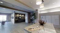 Located at 1971 Willow Loop Way in Knoxville, Tennessee, this single-floor community space offers a polished and contemporary environment designed to welcome residents and visitors alike. The main area greets guests with a prominent granite-topped reception counter, accented by elegant pendant lights that cast a warm, inviting glow throughout the space. Crown molding and neutral-toned walls contribute to a refined architectural aesthetic, while large-format tile flooring adds a clean, modern foundation underfoot. A dedicated refreshment station, set against a striking herringbone-patterned accent wall in dark tones, features cabinetry, a coffee station, and task lighting that creates a cozy, functional nook. Lush potted greenery is thoughtfully placed to bring natural elements indoors, softening the space and enhancing its overall ambiance. A bank of individual mailboxes lines one wall, pointing to the space's practical role within a residential community. Recessed lighting throughout ensures the space remains well-lit, while decorative sculptural accents on the reception counter add artistic character. Overall, the space balances functionality with upscale design, offering a comfortable and informative environment for community members.