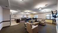 Located in Arden Hills, Minnesota, this single-floor community space offers a thoughtfully designed layout that seamlessly integrates a living room, dining area, and kitchen into one cohesive environment. The living room serves as a welcoming focal point, anchored by a sleek linear gas fireplace set into a large tile surround, with a wall-mounted flat-screen TV above. Comfortable seating — including a sofa and upholstered armchairs — is arranged around a central coffee table, creating an inviting atmosphere for socializing or relaxing. Patterned carpet and warm neutral tones with dark wood wainscoting carry throughout the space, lending a consistent and refined aesthetic. The dining area accommodates multiple groups simultaneously, featuring several high-top tables with granite-style surfaces and dark wood chairs, all illuminated by pendant lighting. Windows along the perimeter bring in natural light and views of the outdoors. The kitchen is equipped with stainless steel appliances — including a full-size refrigerator, range, and microwave — complemented by rich dark cabinetry, granite countertops, and easy-to-clean tile flooring. This well-maintained communal space is ideal for gatherings, meals, and everyday use.