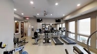 This modern fitness center is thoughtfully designed to support a full workout experience within a clean, well-appointed space. The room features light-toned flooring that provides a bright and open atmosphere, complemented by neutral gray walls and recessed lighting along with a ceiling fan that ensures proper air circulation during workouts.

The cardio equipment lineup is a standout feature, including multiple treadmills, elliptical machines, and a stationary recumbent bike, all equipped with individual digital display consoles. A full-length mirrored wall along the back of the room enhances the sense of space and allows users to monitor their form during exercise. Two wall-mounted flat-screen televisions are positioned for easy viewing from multiple workout stations, adding an entertainment dimension to the fitness experience.

Natural light filters in through large windows along one side of the room, creating an inviting and energizing environment. Additional strength and conditioning equipment is visible throughout the space, offering a well-rounded selection for users of varying fitness levels. Safety features such as a fire extinguisher and clearly marked emergency exit signage are also present. The overall ambiance is clean, functional, and professional, making this fitness center a practical and welcoming amenity.