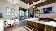 This single-floor amenity space is designed with a contemporary aesthetic, blending upscale finishes with versatile social zones. At its center sits a striking bar island clad in ribbed brass paneling and topped with a crisp white quartz countertop, complemented by a full service wall featuring mounted displays, open shelving, and a beer tap. A coffered ceiling with deep dark wood panels and globe pendant lighting anchors the bar area, lending warmth and architectural depth.

Adjacent to the bar, a banquette dining nook provides an intimate setting with upholstered booth seating and a round white table, bathed in natural light filtering through steel-framed interior windows. The lounge area transitions seamlessly into a sun-drenched billiards and social room encased in expansive floor-to-ceiling windows, offering sweeping urban views and direct outdoor access. High ceilings amplify the sense of openness throughout this wing.

A separate lounge retreat features a curated gallery wall, large-screen television, and plush seating arranged around a geometric area rug. Rich wood-plank flooring runs throughout, tying each zone together. The overall ambiance is polished yet welcoming, designed to encourage both relaxed gatherings and lively social interaction.